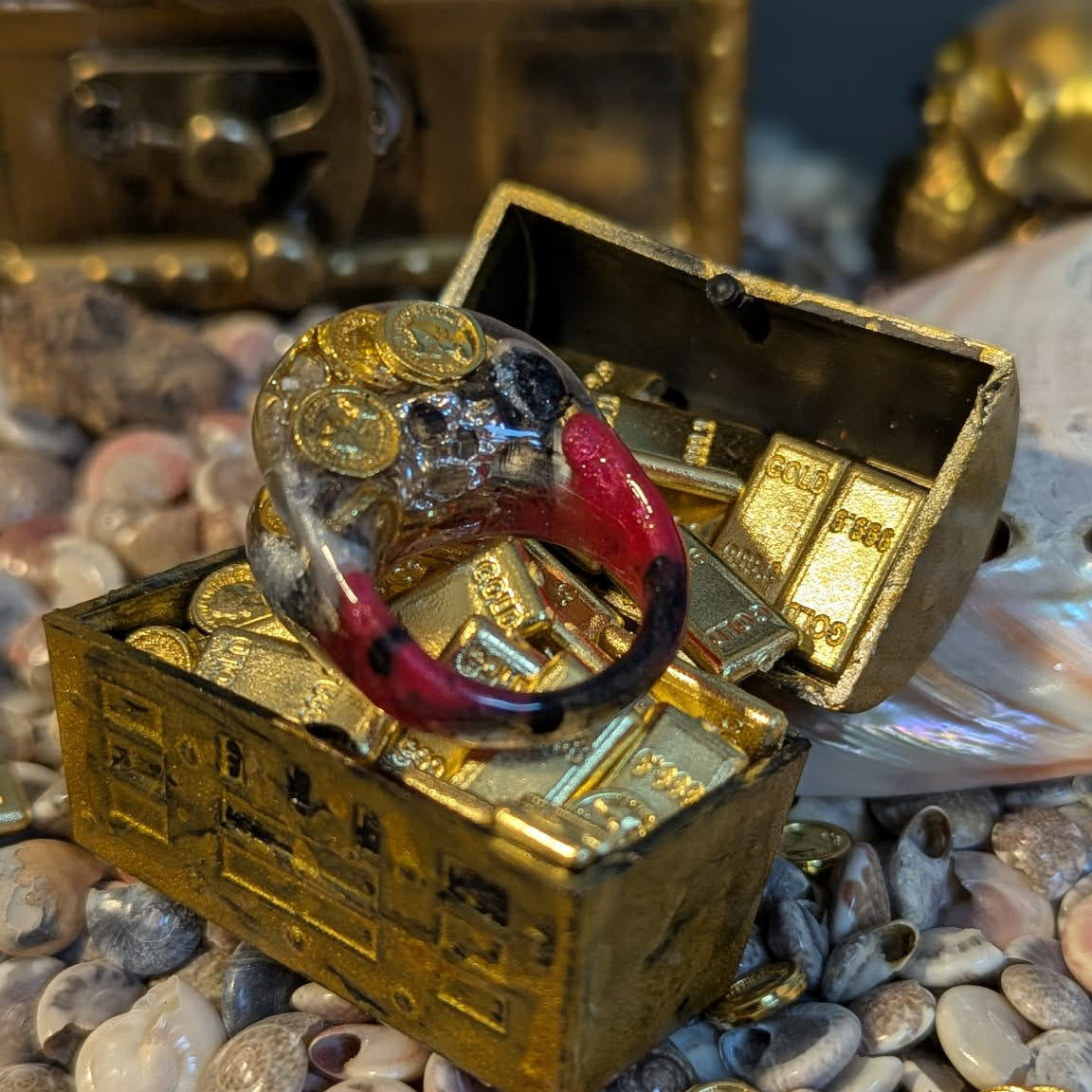 Red and black ring with coins on a miniature gold treasure chest surrounded by pebbles and coins.